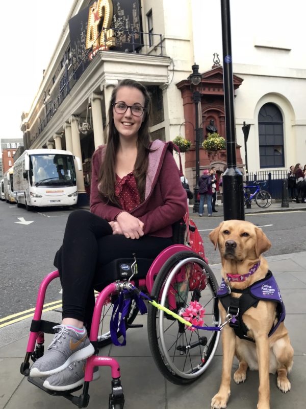 Lauren is sitting in a pink wheelchair in front of a sign for '42nd Street The Musical'. Her fox red assistance dog is sitting to the left. 