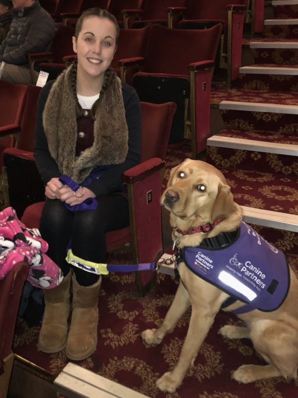 Lauren is sitting inside a London theatre. Her fox red assistance dog is sitting to the left. 