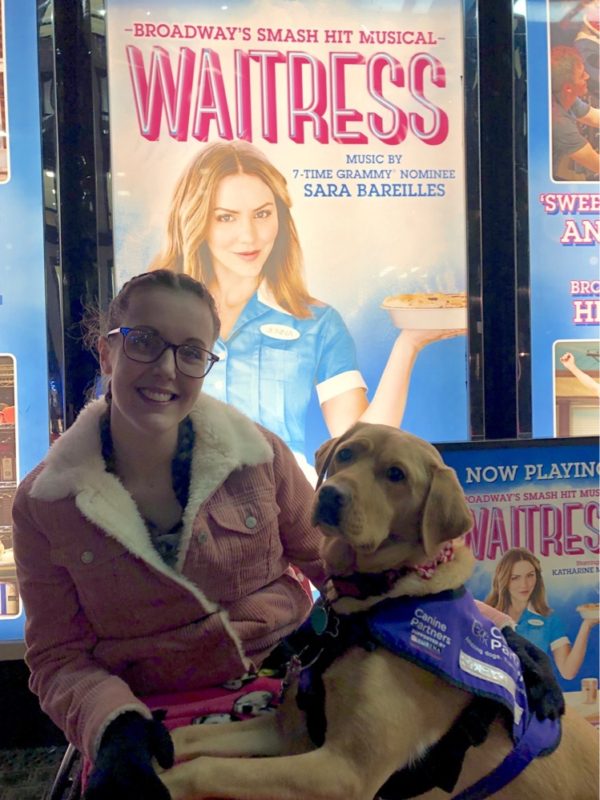 Lauren is sitting in a pink wheelchair in front of a sign for 'Waitress The Musical'. Her fox red assistance dog is sitting to the left. 