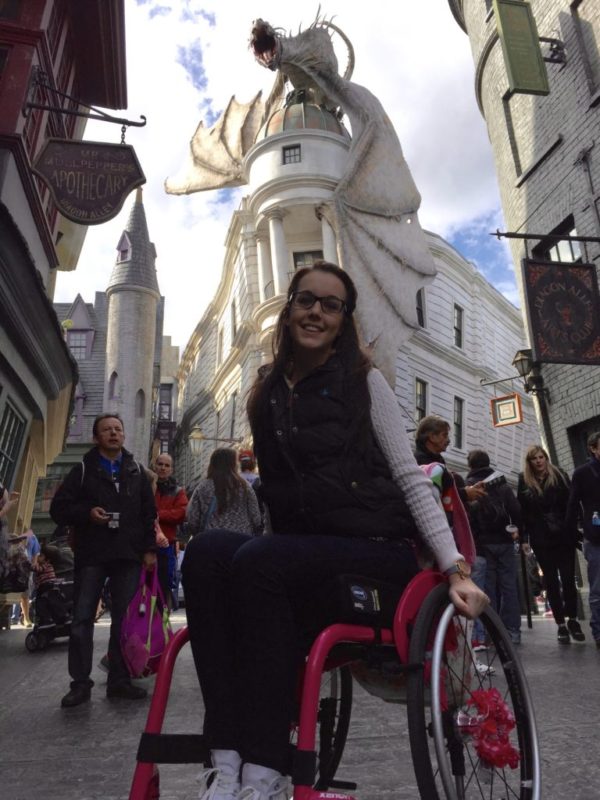 Lauren is wearing a black body warmer whilst sitting in a pink wheelchair in front of Gringotts Bank at Universal Studios. 