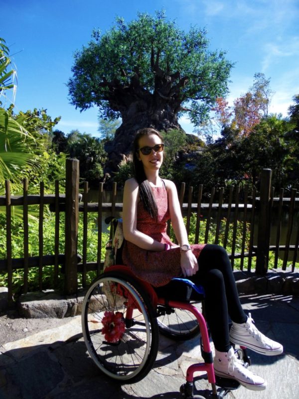 Lauren is wearing a red dress whilst sitting in a pink wheelchair in front of the Tree of Life at Disney's Animal Kingdom.