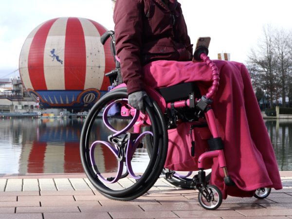 Purple Loopwheels attached to a pink manual wheelchair. You can only see the users legs, which are wrapped in a matching fleece blanket.