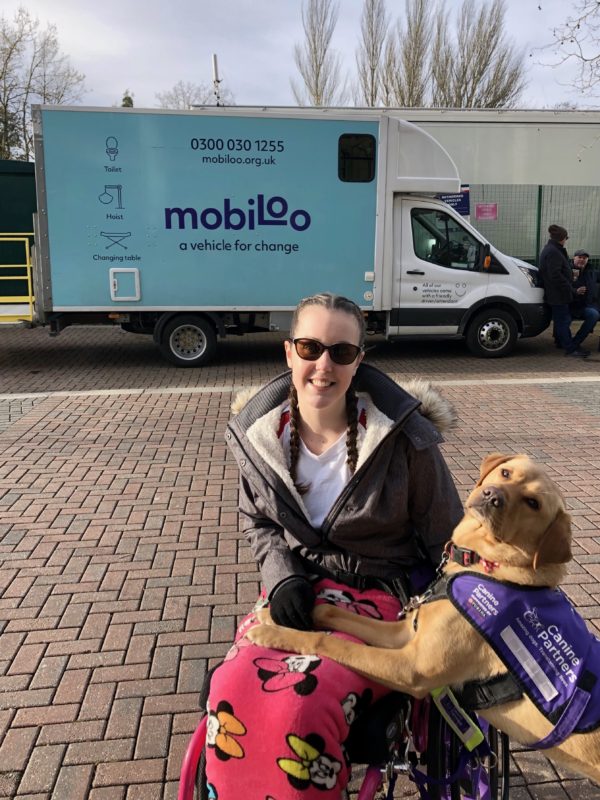 Lauren is sitting in a bright pink wheelchair with her fox red assistance dog stood to her left-hand side. They are both sat in front of a Mobiloo van.