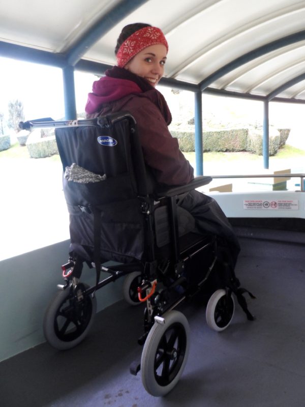 Lauren is wearing a purple coat, scarf and hat whilst sitting in a bulky black wheelchair. She is looking over her shoulder and smiling directly at the camera.