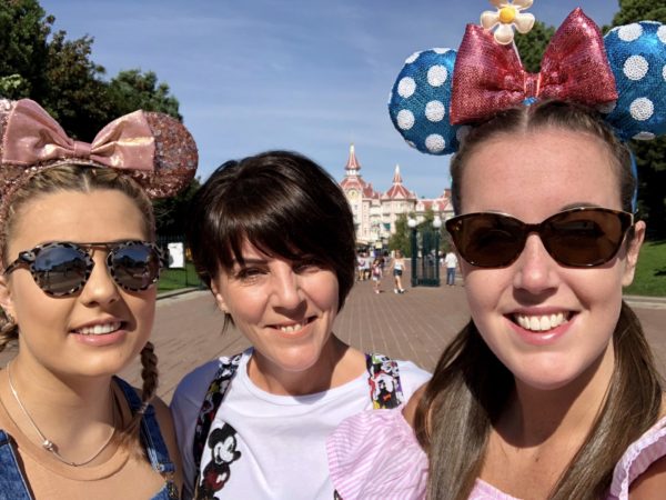 Lauren is standing in front of the Disneyland Hotel in Paris, alongside her mum and best friend. They are all wearing sparkly Minnie Mouse ears whilst smiling directly at the camera.