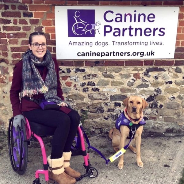 Lauren is sitting in a bright pink wheelchair and smiling directly at the camera. Her fox red assistance dog is sat to her left-hand side. They are both sat in front of a sign for the UK charity Canine Partners. 