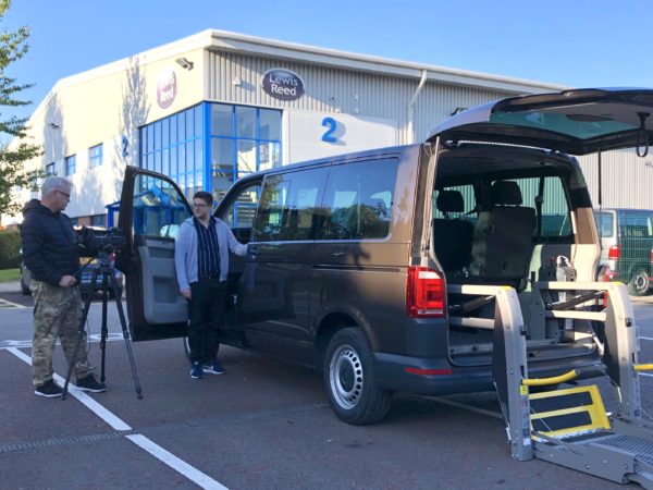 Felix is stood by the driver's seat of a dark grey VW T6 Transporter, which is parked outside of the Lewis Reed office. A cameraman is to his left-hand side filming the process.