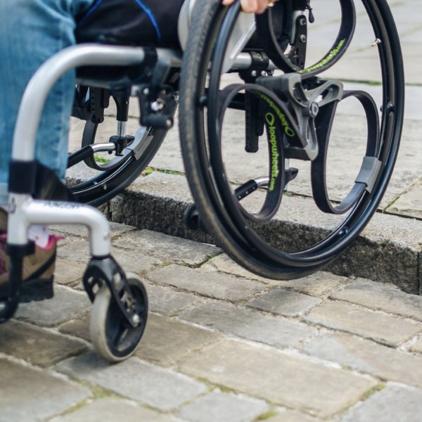 Black Loopwheels attached to a silver manual wheelchair. You can only see the users hand on one of the black push rims, as the wheels roll down a kerb.