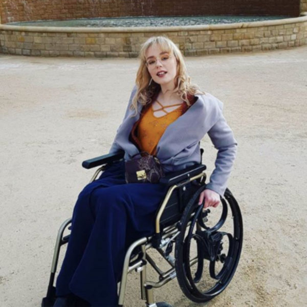 Lori is sitting in a gold wheelchair with black Loopwheels, whilst using a navy blanket to keep warm and smiling directly at the camera.