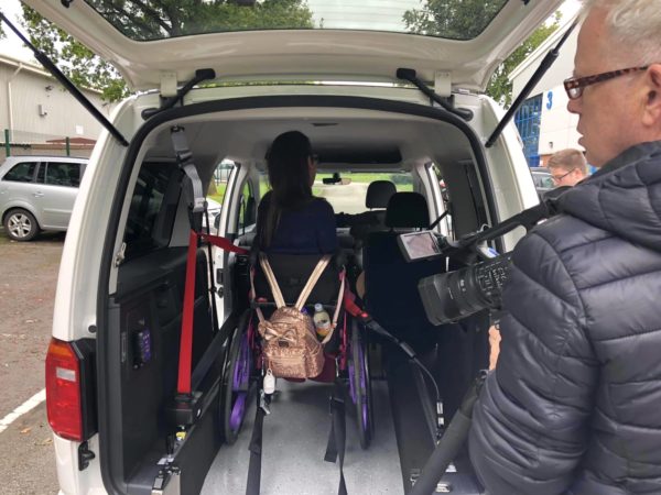 Lauren is sitting in a bright pink wheelchair, whilst being strapped into a white VW Caddy Maxi. A cameraman is to her right-hand side filming the process.