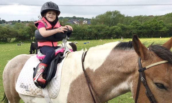 A little girl is sat on a horse with a specialised saddle that supports her spine. She is wearing pink riding gear and smiling directly at the camera. 