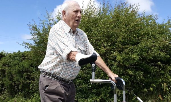 An elderly gentleman with an amputated arm is using an adapted walking frame, which has a raised handlebar on one side to support his amputated arm.