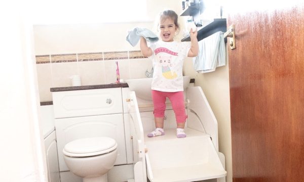 A young girl with dwarfism is stood next to a bathroom sink with the help of a specially adapted step stool! She is holding a blue washcloth and smiling directly at the camera. 