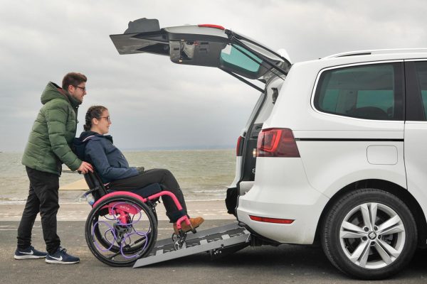 Lauren is sitting in a bright pink wheelchair that is being pushed up the access ramp of a white Seat Alhambra by her carer Felix.