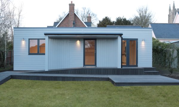 A small grey annexe is situated at the bottom of a garden, opposite a family home. 