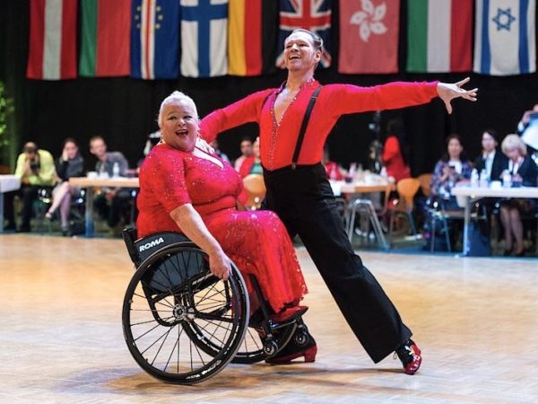 Two Para Dance athletes performing together (one wheelchair user and one able-bodied). They are both wearing red sparkly costumes.