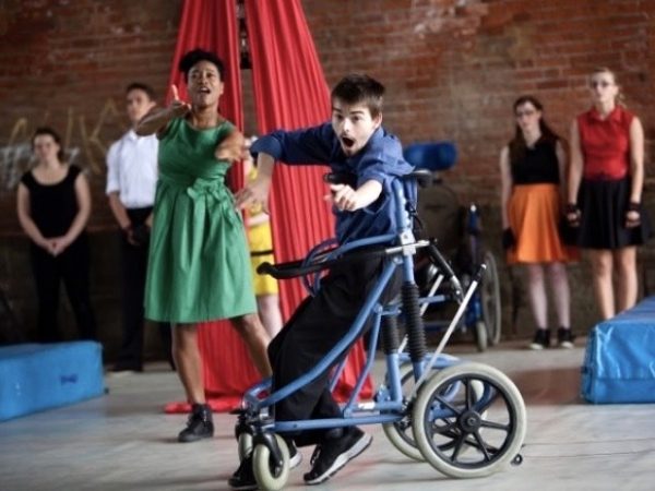 A young boy using a blue walking frame is dancing during an inclusive class. Classmates are cheering in the background.