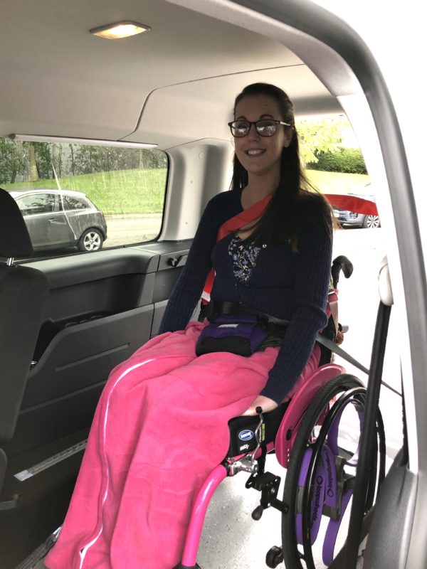 Lauren is sitting in a bright pink wheelchair that is strapped into a white Volkswagen Caddy Maxi. She is smiling directly at the camera and wearing a navy cardigan with pink wheelchair blanket. 
