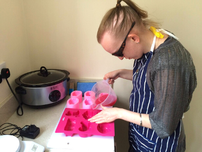 Alexandra Negoita is a young woman wearing dark glasses and an apron. She is pouring soap into pink soap moulds.