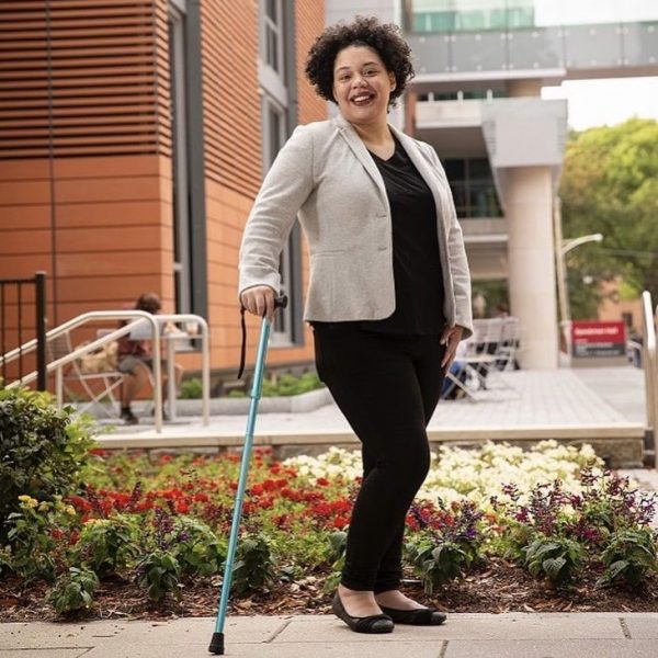 A picture of a woman, Natasha Graves, standing with a cane. She is looking at the camera and smiling.