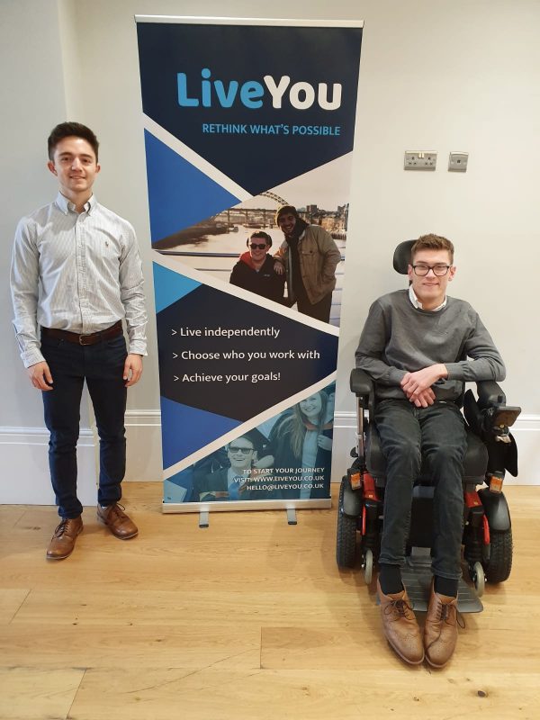 Daniel (right) and Sam (left), the founder and co-founder of LiveYou, are on either side of a banner stand. Sam is standing and Daniel is in a red and black powerchair, and they are smiling at the camera. The text on the banner stand reads: LiveYou. Rethink what's possible.  Live independently, choose who you work with, achieve your goals!